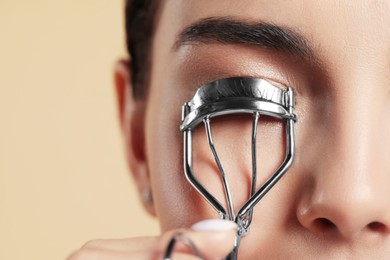 Young woman using eyelash curler on beige background, closeup