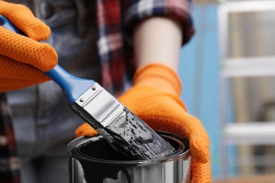 Photo of Woman dipping brush into can of black paint indoors, closeup