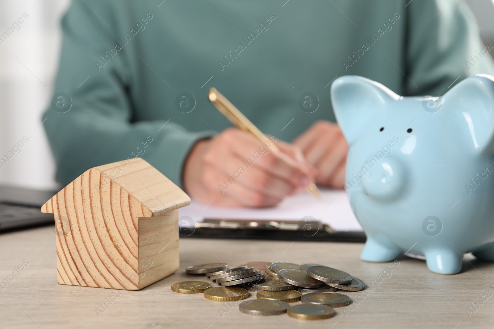 Photo of Woman planning budget at wooden table, focus on house model, piggy bank and coins