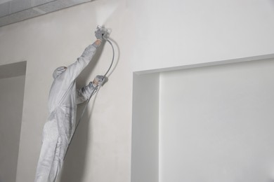 Photo of Decorator in protective overalls painting wall with spray gun indoors