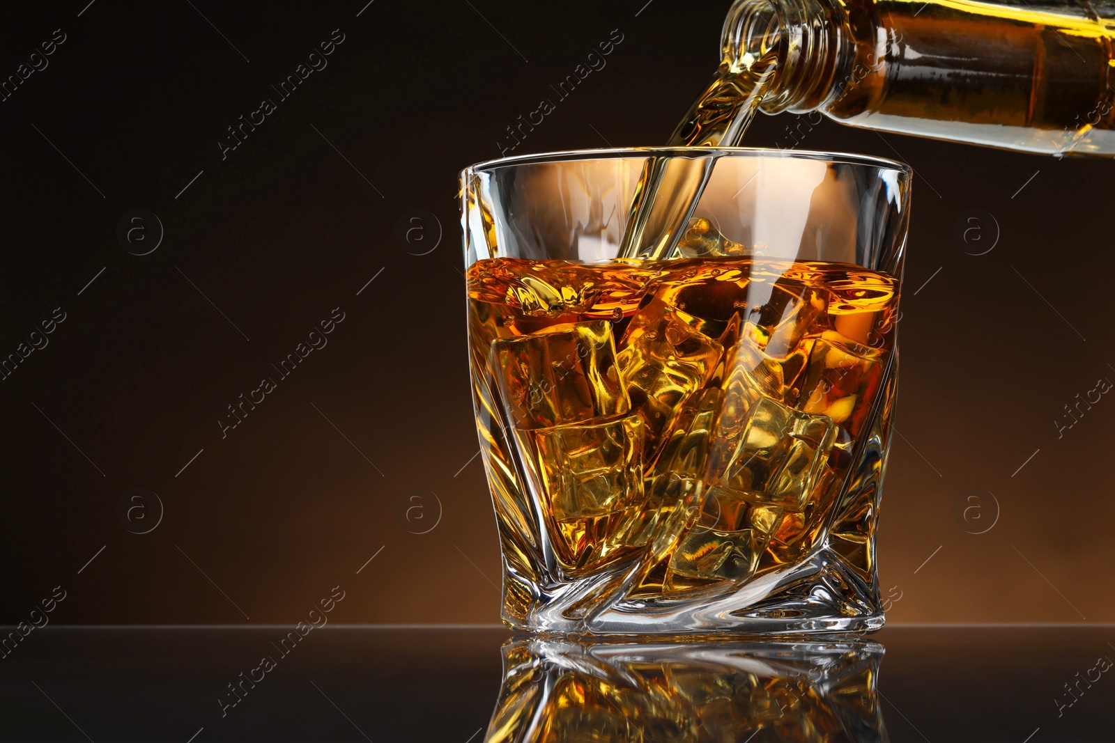 Photo of Pouring whiskey from bottle into glass with ice cubes at table against brown background, space for text