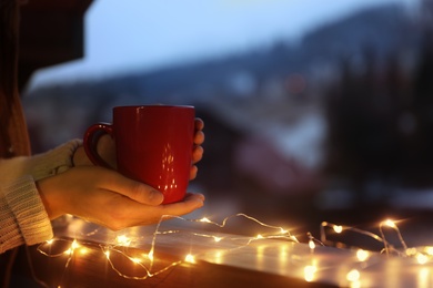 Woman holding cup of hot beverage on balcony decorated with Christmas lights, closeup with space for text. Winter evening