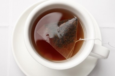 Tea bag in cup with hot drink on white tiled table, top view