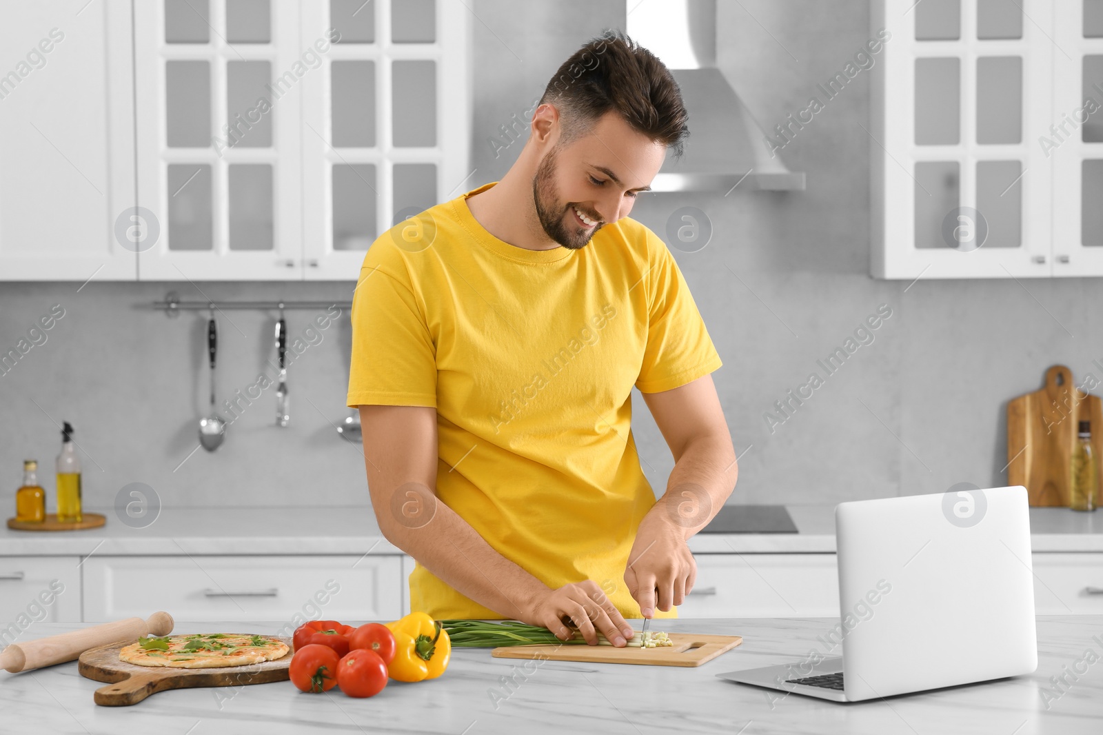 Photo of Man watching cooking online course on laptop while cutting scallion in kitchen. Time for hobby