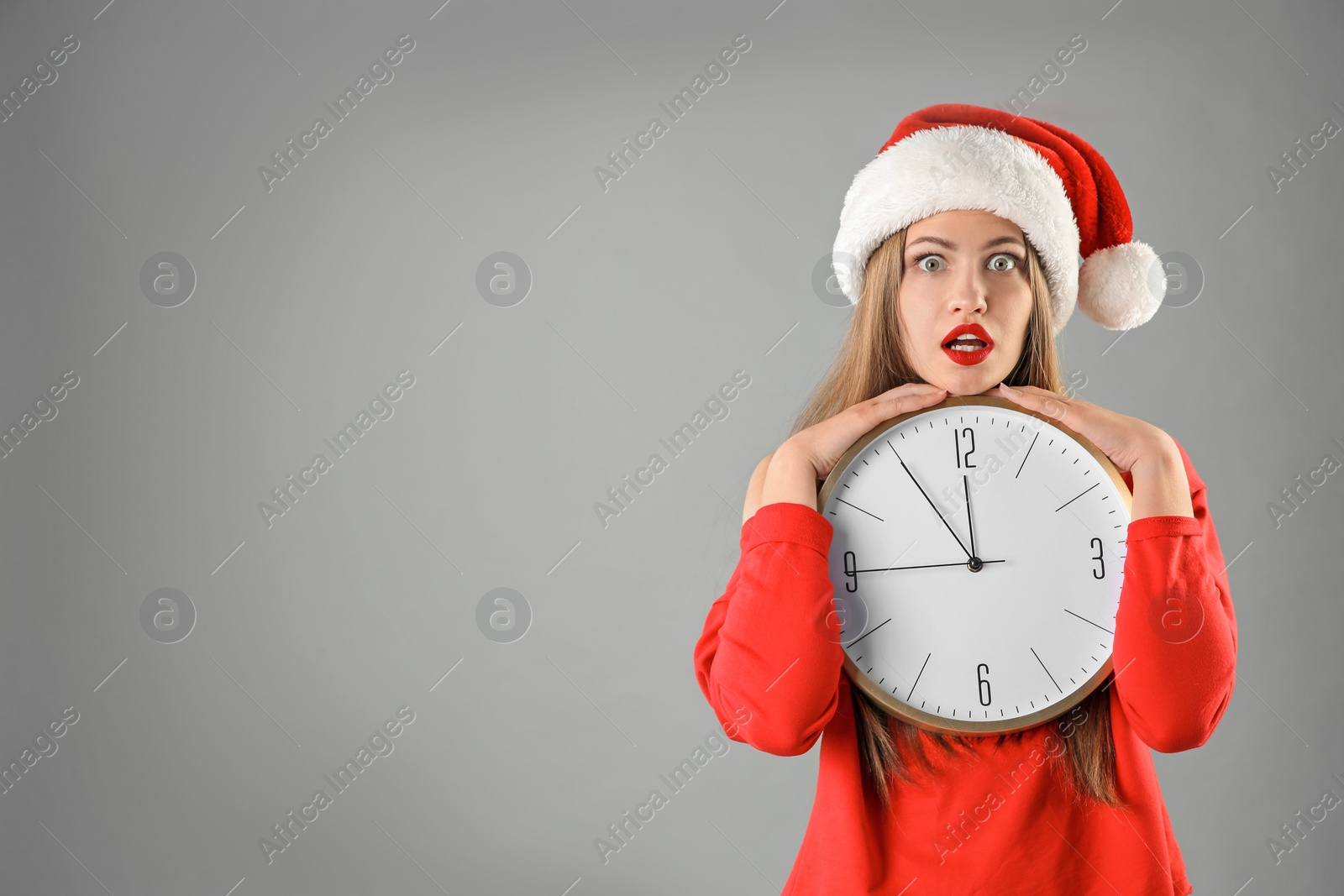 Photo of Young beautiful woman in Santa hat holding big clock on grey background. Christmas celebration