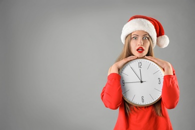 Photo of Young beautiful woman in Santa hat holding big clock on grey background. Christmas celebration