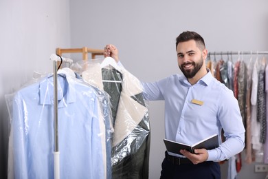 Dry-cleaning service. Happy worker holding hanger with coat in plastic bag indoors