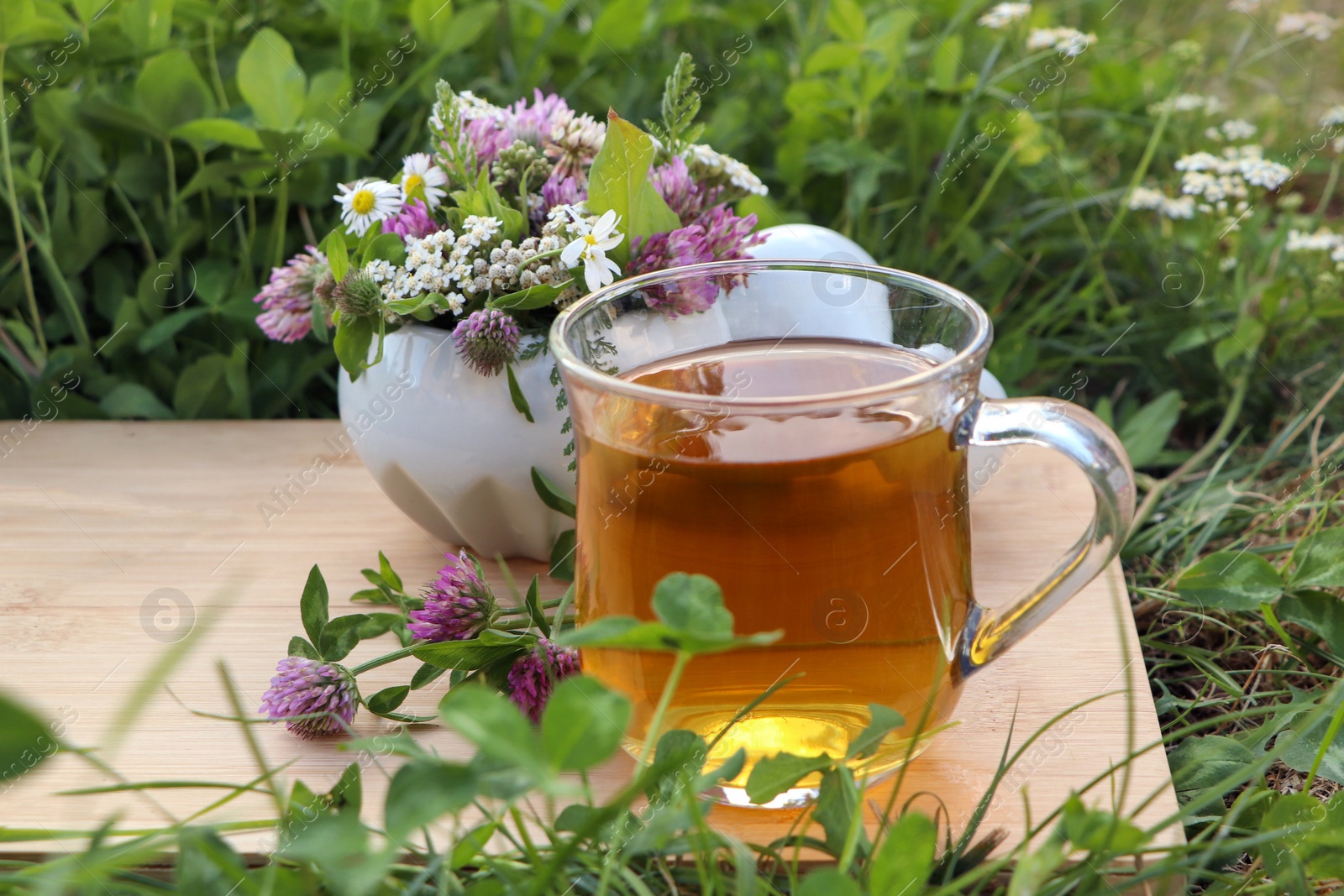 Photo of Cup of aromatic herbal tea, pestle and ceramic mortar with different wildflowers on green grass outdoors