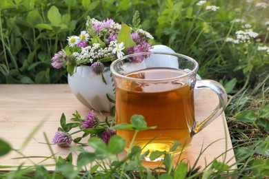 Photo of Cup of aromatic herbal tea, pestle and ceramic mortar with different wildflowers on green grass outdoors