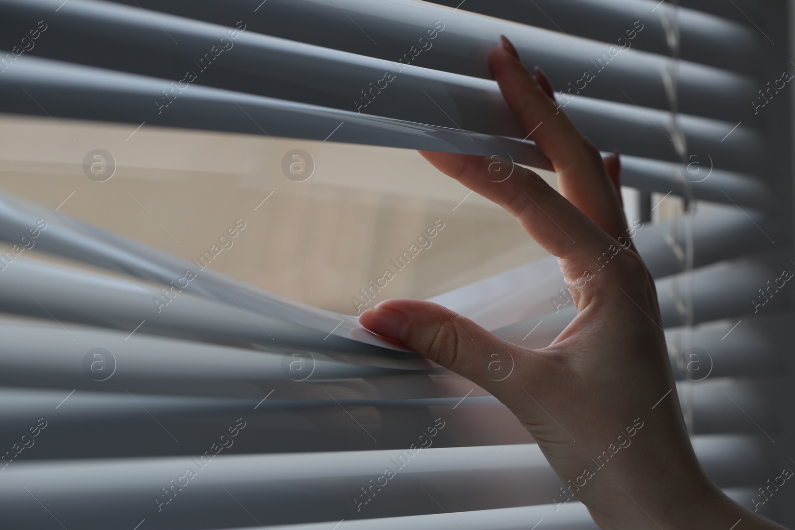 Photo of Woman separating slats of white blinds indoors, closeup