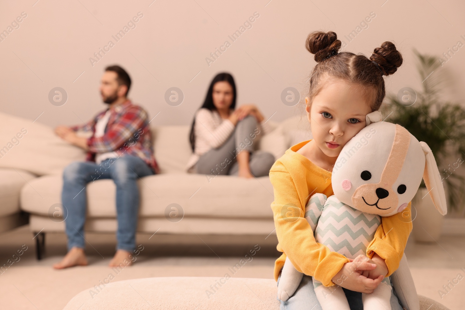 Photo of Sad little girl with toy and her arguing parents on sofa in living room