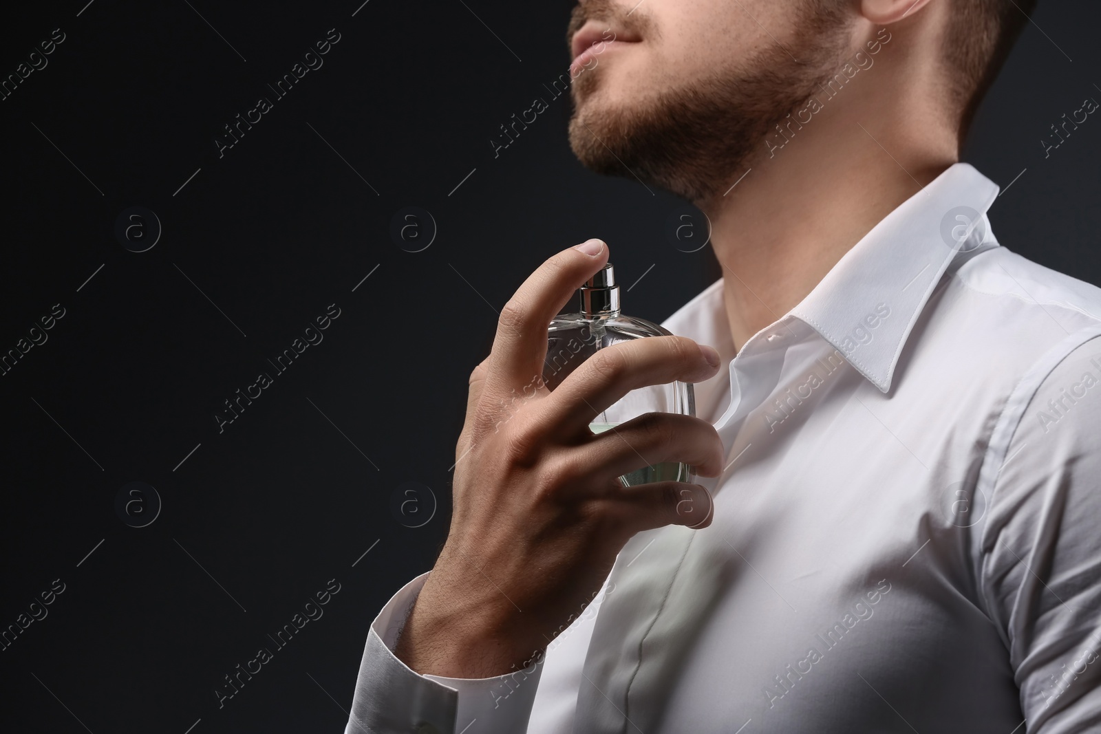 Photo of Handsome man in shirt using perfume on dark background, closeup