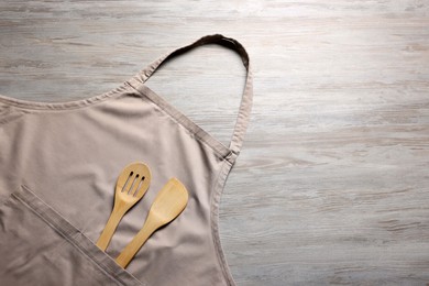 Photo of Stylish beige apron, spatula and slotted spoon on light wooden table, top view. Space for text