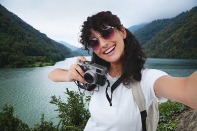 Beautiful woman in sunglasses with camera taking selfie near river in mountains