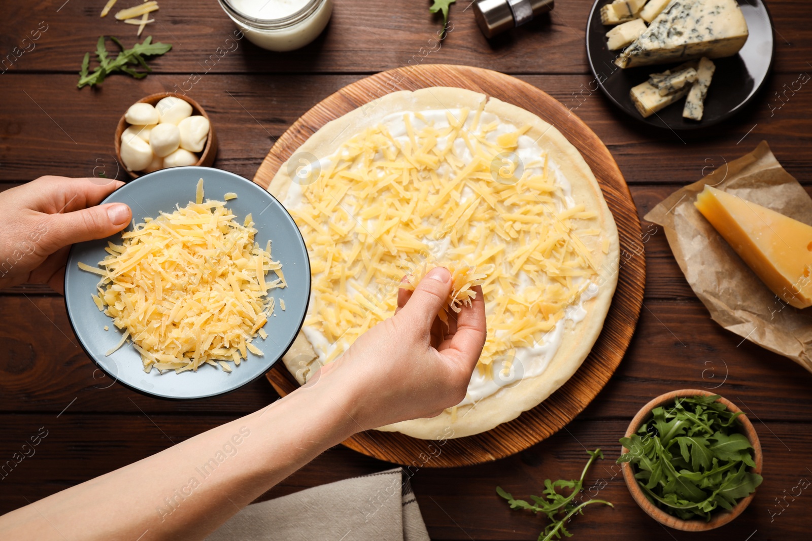 Photo of Woman adding grated cheese to unbaked pizza on wooden table, top view