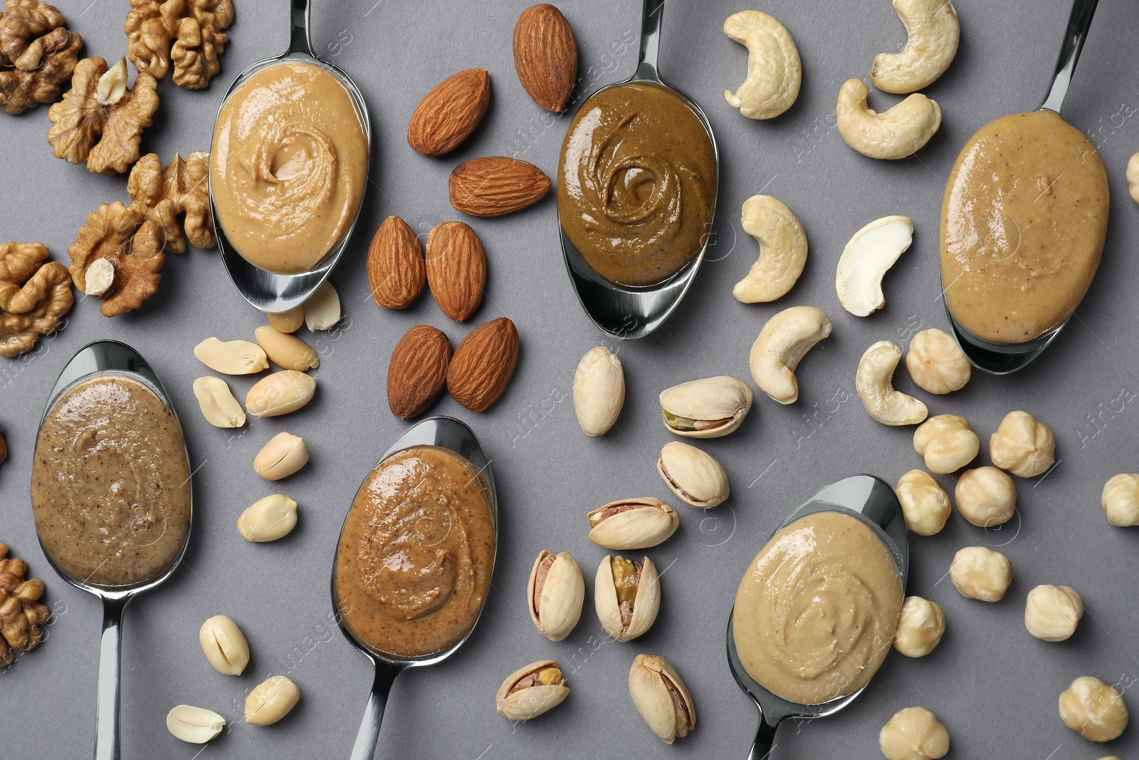 Photo of Tasty nut butters in spoons and raw nuts on gray table, flat lay