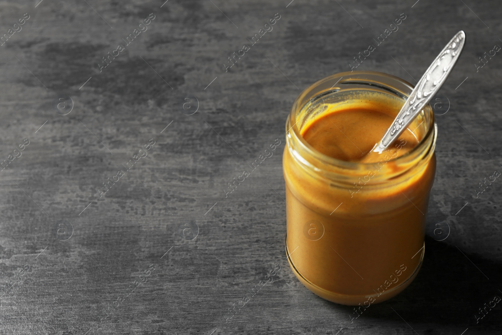 Photo of Spicy mustard and spoon in glass jar on dark grey table. Space for text