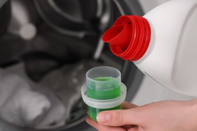 Photo of Woman pouring fabric softener from bottle into cap near washing machine, closeup