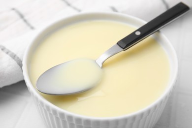 Photo of Bowl with condensed milk and spoon on white table, closeup