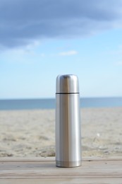 Metallic thermos with hot drink on wooden surface near sea