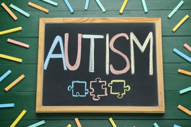 Photo of Blackboard with word Autism and drawn jigsaw puzzle pieces surrounded by colorful chalks on green wooden background, flat lay