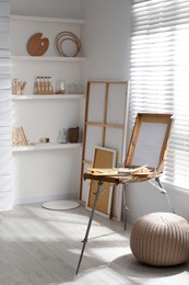 Photo of Modern studio interior with artist's workplace and wooden easel near large window