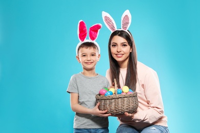 Photo of Mother and son in bunny ears headbands with basket of Easter eggs on color background