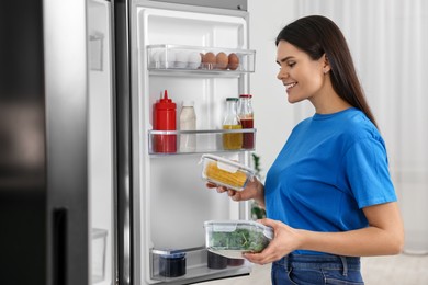 Photo of Young woman taking containers with vegetables out of refrigerator indoors