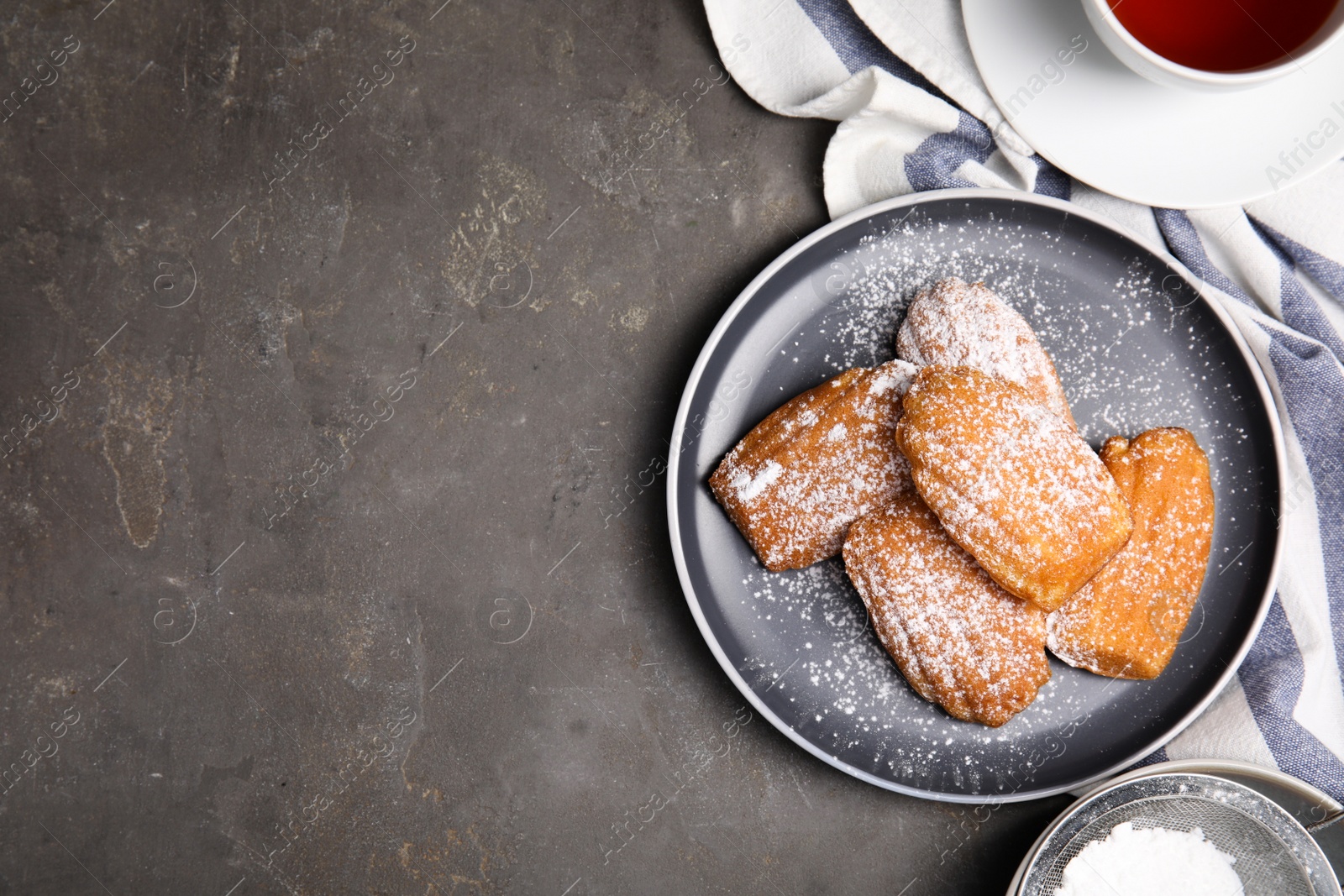 Photo of Delicious madeleine cakes with powdered sugar and tea on grey table, flat lay. Space for text