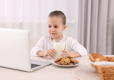 Photo of Little girl using laptop while having breakfast at table indoors. Internet addiction