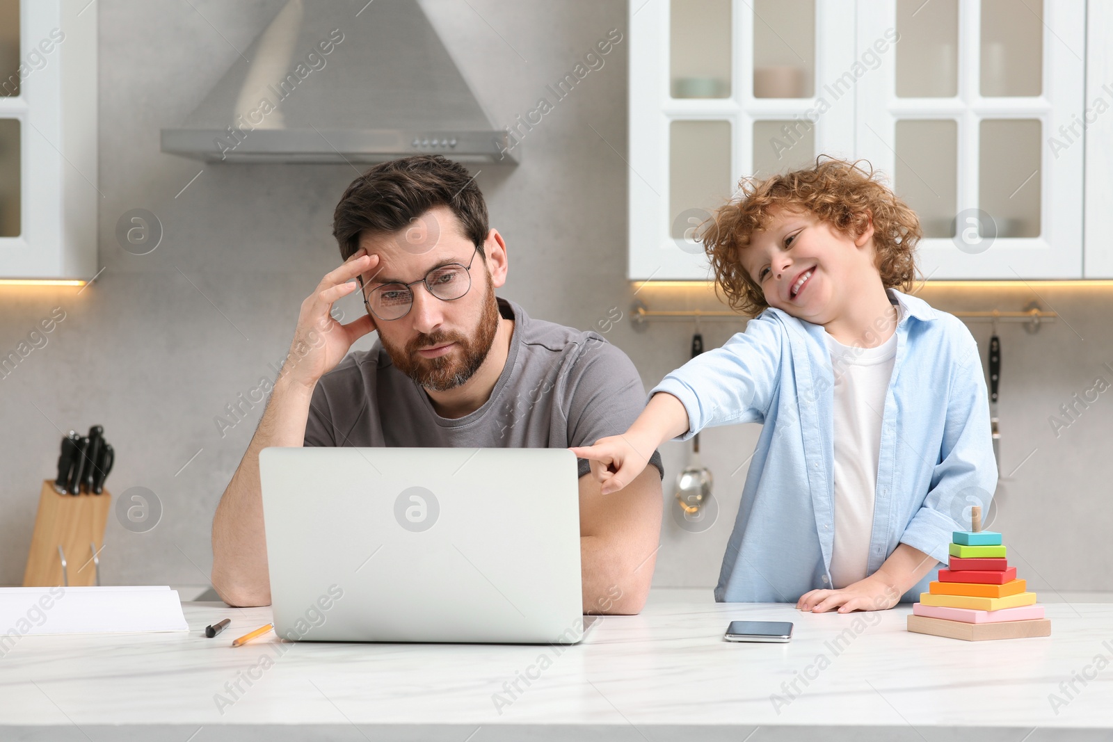 Photo of Little boy bothering father while he working remotely at home. Man with laptop and his child at desk in kitchen