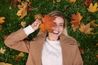 Photo of Beautiful woman lying on grass and covering eye with autumn leaf, top view