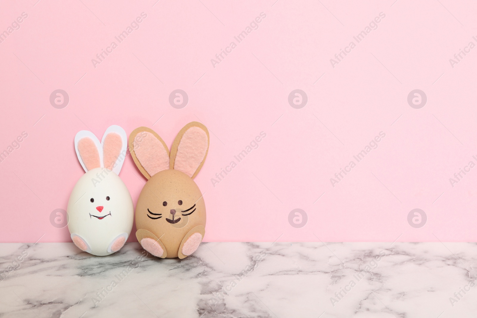 Photo of Eggs as cute bunnies on white marble table against pink background, space for text. Easter celebration