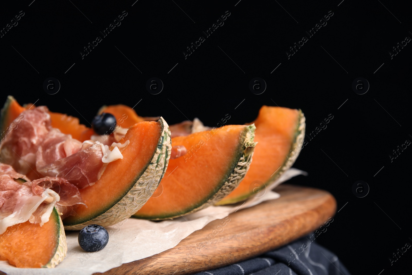 Photo of Wooden board with melon, prosciutto and blueberries on table against black background