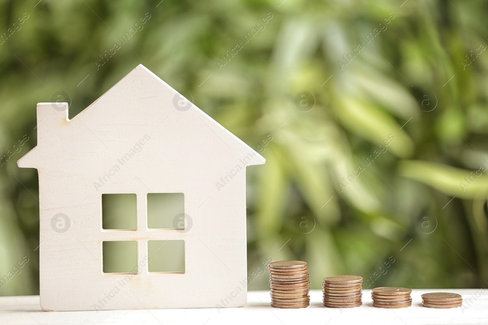 Photo of House model and coins on white table against blurred green background. Space for text