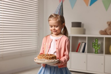 Photo of Birthday celebration. Cute little girl in party hat holding tasty cake with burning candles at home