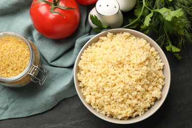 Photo of Delicious bulgur in bowl, dill, parsley and tomato on black table, top view