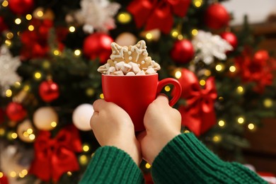 Photo of Woman holding cup of delicious cocoa with marshmallows and gingerbread cookie near Christmas tree, closeup