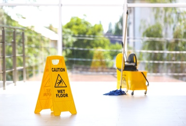 Photo of Safety sign with phrase Caution wet floor and mop bucket, indoors. Cleaning service