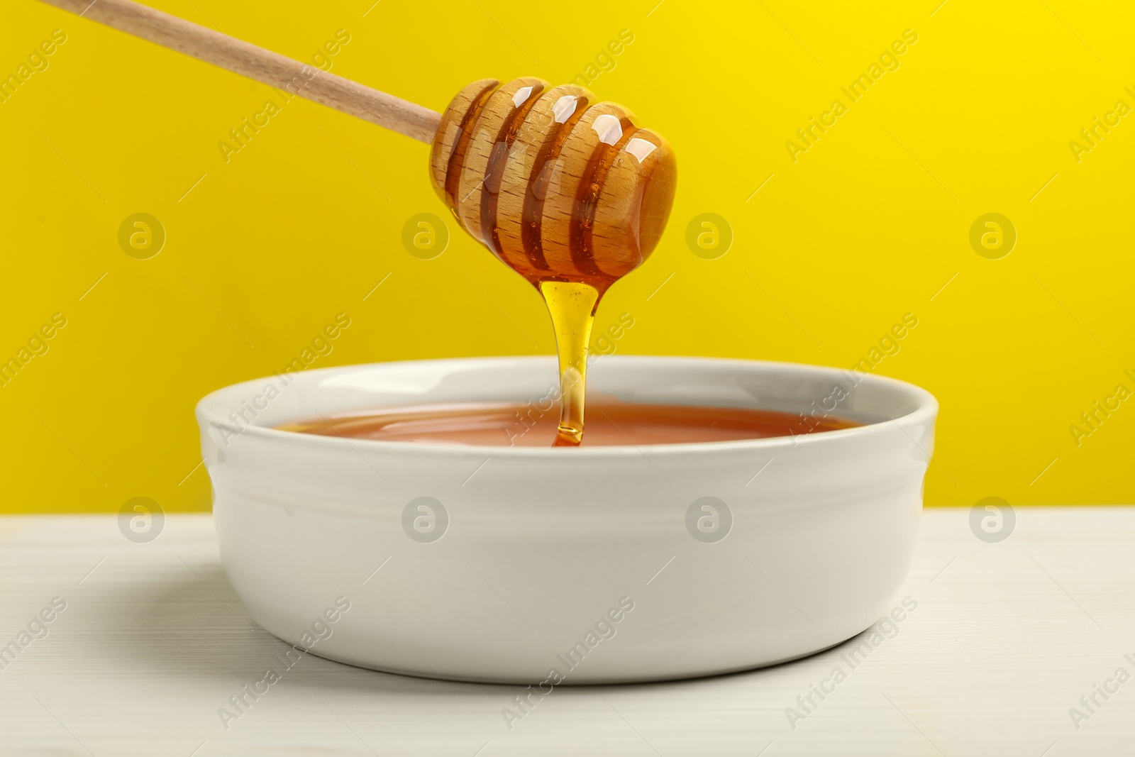 Photo of Pouring tasty honey from dipper into bowl at white wooden table against yellow background, closeup