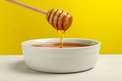 Photo of Pouring tasty honey from dipper into bowl at white wooden table against yellow background, closeup