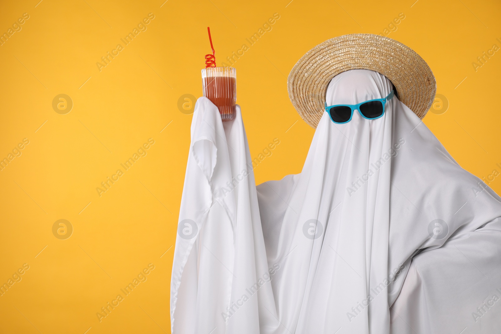 Photo of Person in ghost costume, sunglasses and straw hat holding glass of drink on yellow background, space for text