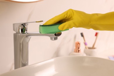 Photo of Woman in gloves cleaning faucet of bathroom sink with sponge, closeup