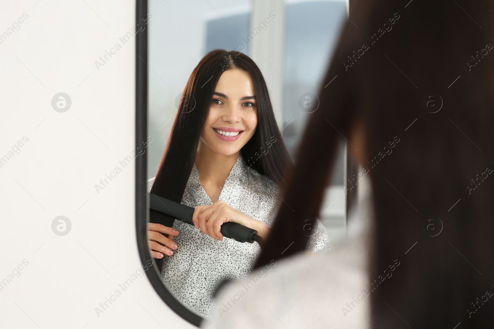 Photo of Beautiful happy woman using hair iron near mirror indoors