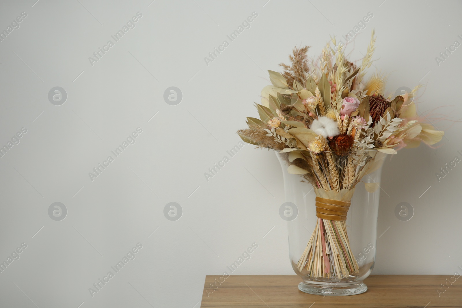 Photo of Beautiful dried flower bouquet in glass vase on wooden table near white wall. Space for text