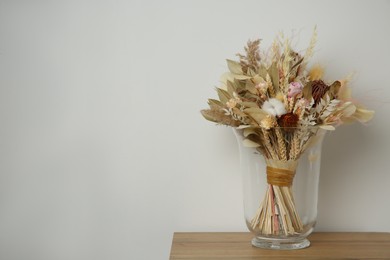 Beautiful dried flower bouquet in glass vase on wooden table near white wall. Space for text