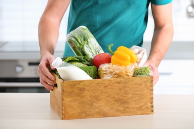 Man with wooden crate full of products at table in kitchen, closeup. Food delivery service