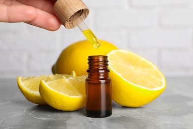 Photo of Woman dripping essential oil into bottle and lemons on grey marble table, closeup
