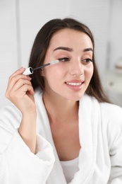 Attractive young woman applying oil onto her eyelashes indoors