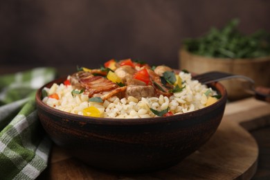 Cooked bulgur with vegetables, fried bacon and mushrooms in bowl on wooden board, closeup